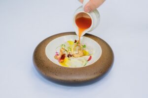Sauce in a small white jug being poured onto a delicate arrangement of vegetables in a round white dish with brown border